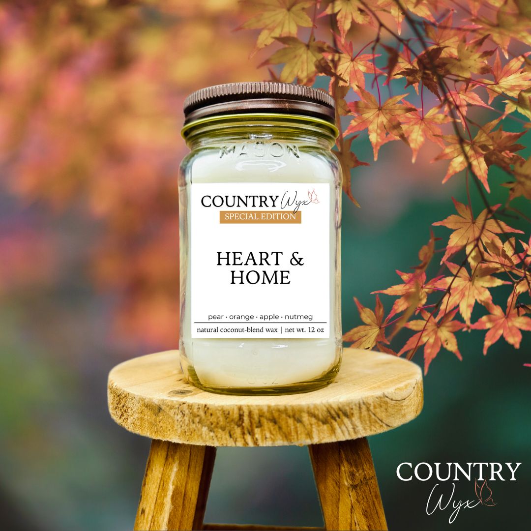 Candle jar labeled &#39;Heart &amp; Home&#39; by Country Wyx on a wooden stool with autumn leaves in the background.