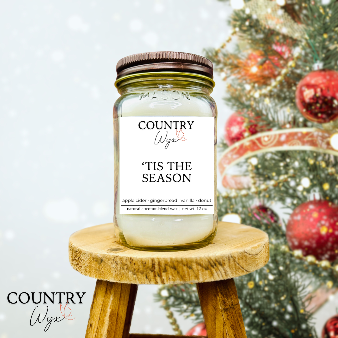 'Tis The Season 16oz candle by Country Wyx on a wooden stool with a blurred Christmas tree and ornaments in the background.