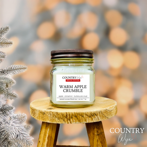 Warm Apple Crumble 8oz candle by Country Wyx on a wooden stool with a frosted pine tree holiday background