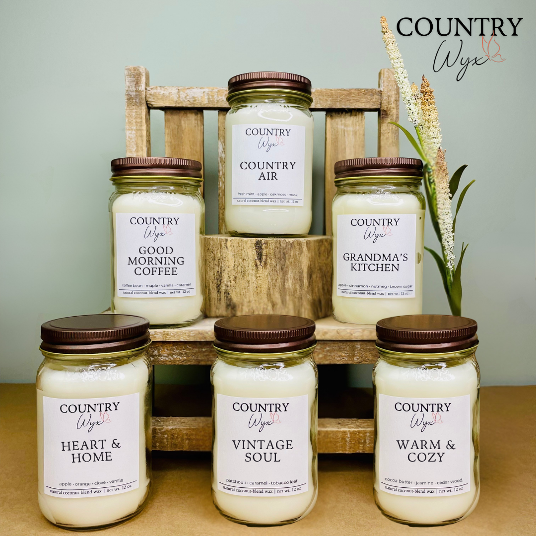 Country Wyx 16oz candles displayed on a rustic wooden shelf with farmhouse-inspired decor, featuring Country Air, Good Morning Coffee, Grandma’s Kitchen, Heart &amp; Home, Vintage Soul, and Warm &amp; Cozy