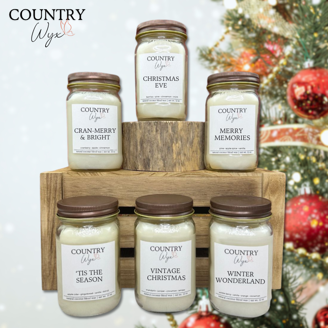 Winter Collection of Country Wyx 16oz candles with holiday-themed labels on a wooden stand against a blurred Christmas tree with ornaments background.